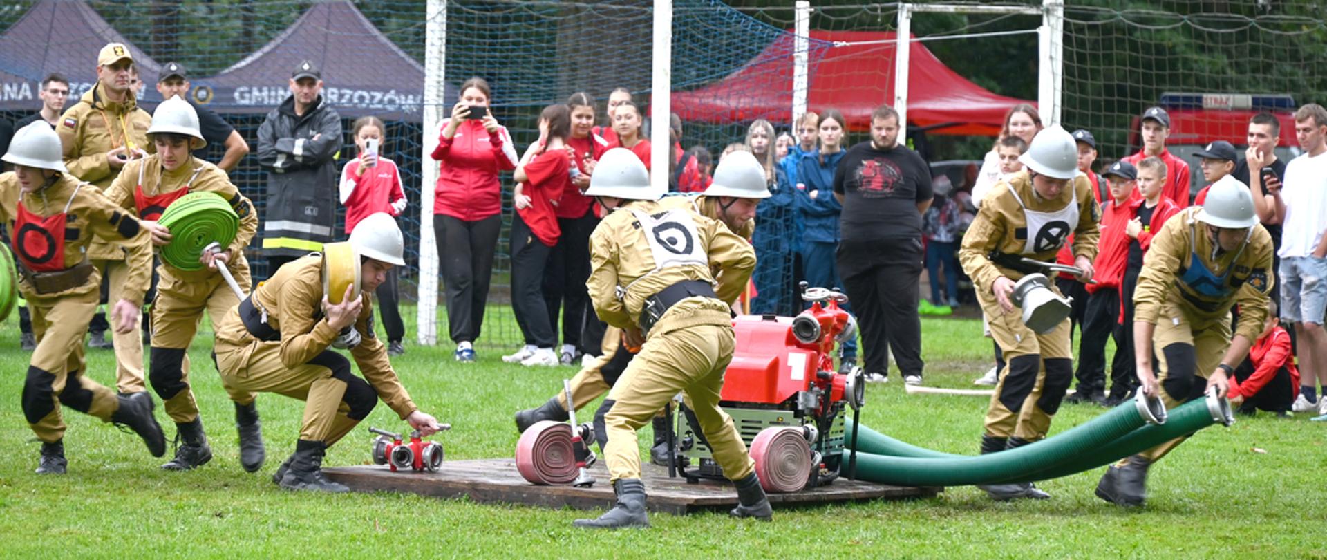 Kolorowa fotografia wykonana w deszczowy dzień na stadionie piłkarskim. Przedstawia strażaków OSP w Powiatowych Zawodach Sportowo Pożarniczych podczas ćwiczenia bojowego. Strażacy ubrani w ubrania służbowe z hełmami rozwijają węże z podestu. W tle kibice obserwujący start drużyny. 