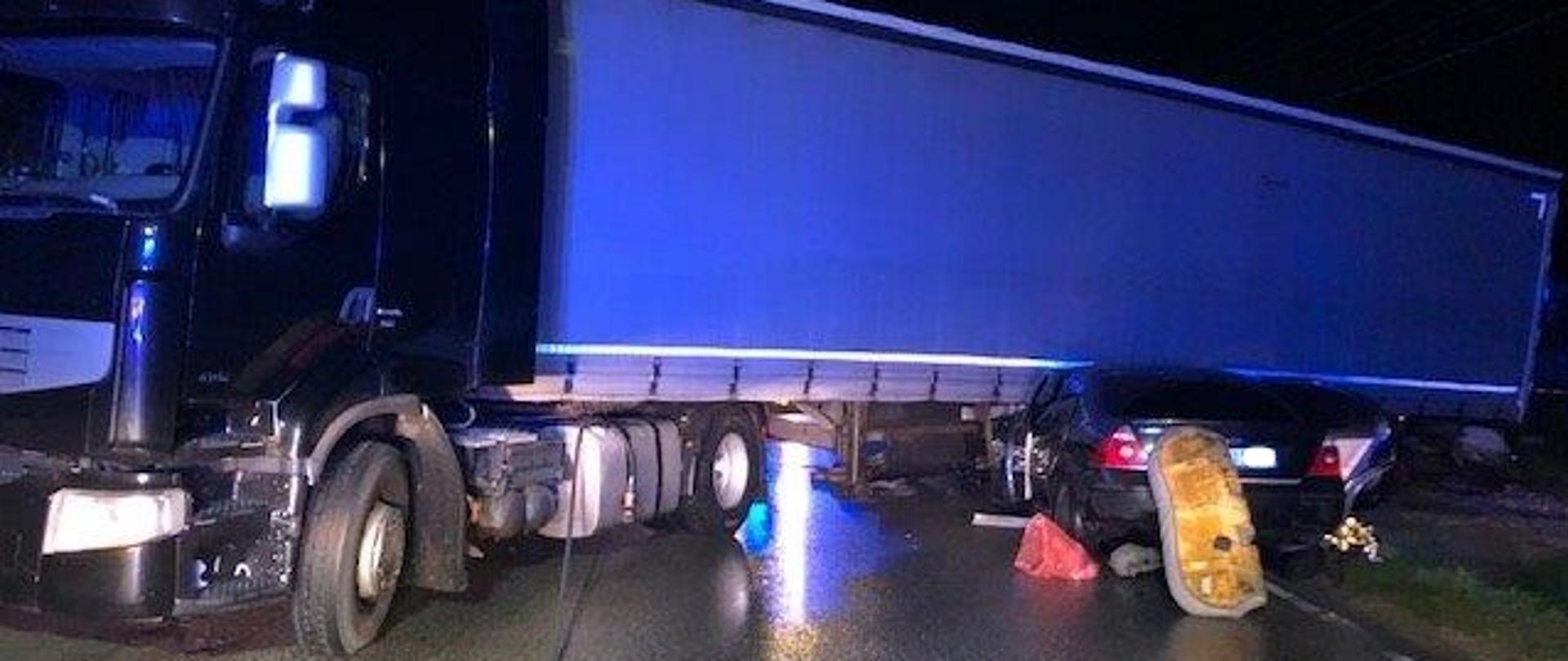 Zdjęcie wykonane w porze nocnej. Na jezdni znajduje się samochód osobowy marki Ford Mondeo, który wjechał pod naczepę samochodu ciężarowego marki Reanault. Samochód ciężarowy ustawiony jest w poprzek drogi, prawdopodobnie wykonując manewr cofania, natomiast pojazd osobowy znajduje się częściowo pod naczepą. Ciągnik siodłowy pojazdu w kolorze czarnym, naczepa w kolorze niebieskim. Pojazd osobowy w kolorze czarnym. 