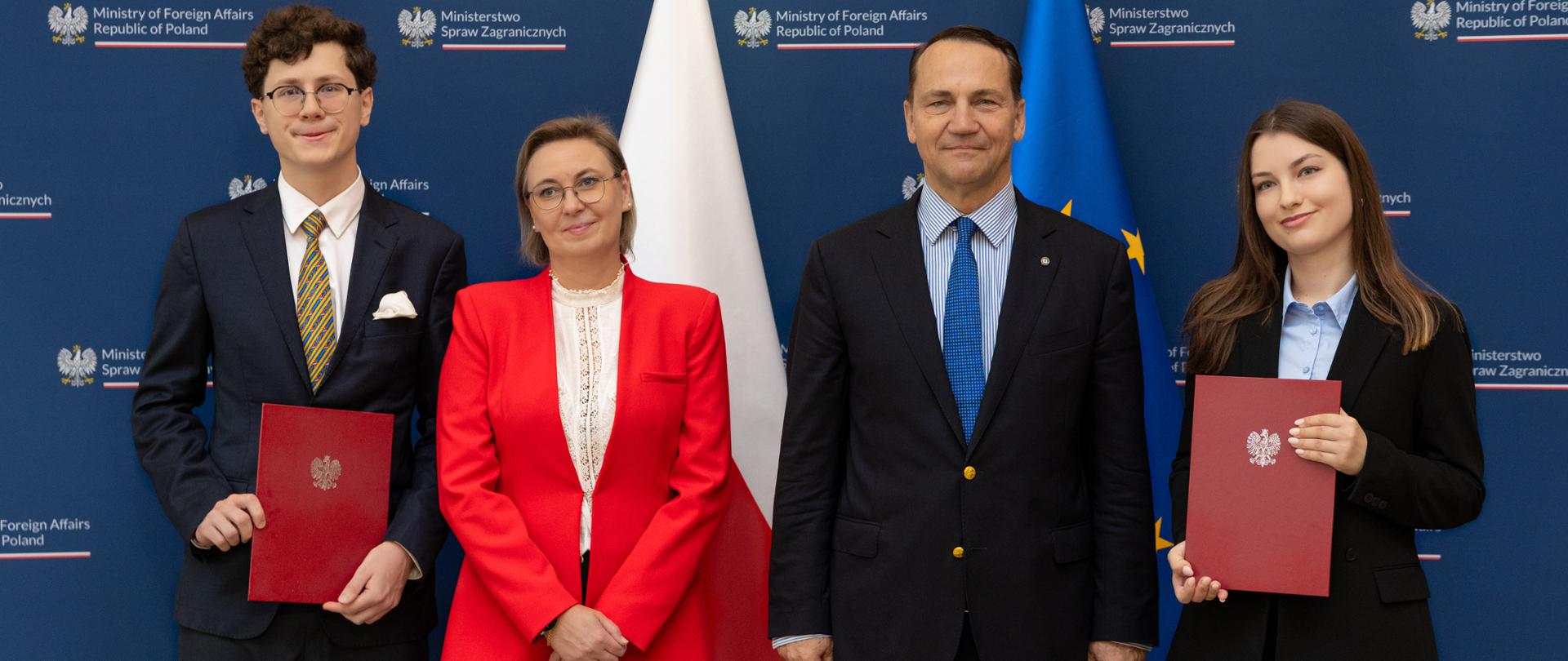 There are four people in the photo. They are standing side by side and looking straight ahead. Two of them are holding red folders. Behind them are the Polish and EU flags. 