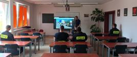 Zdjęcie przedstawia strażaków w sali szkoleniowej, którzy słuchają policjanta prowadzącego szkolenie