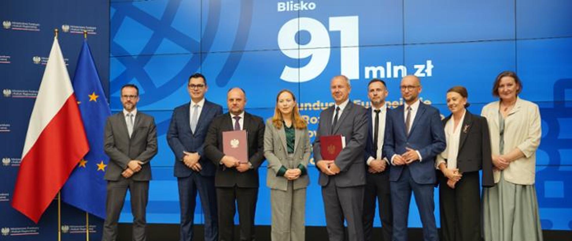 In the photo, Minister Katarzyna Pełczyńska-Nałęcz stands on stage surrounded by other conference participants. The Polish and EU flags are visible in the background