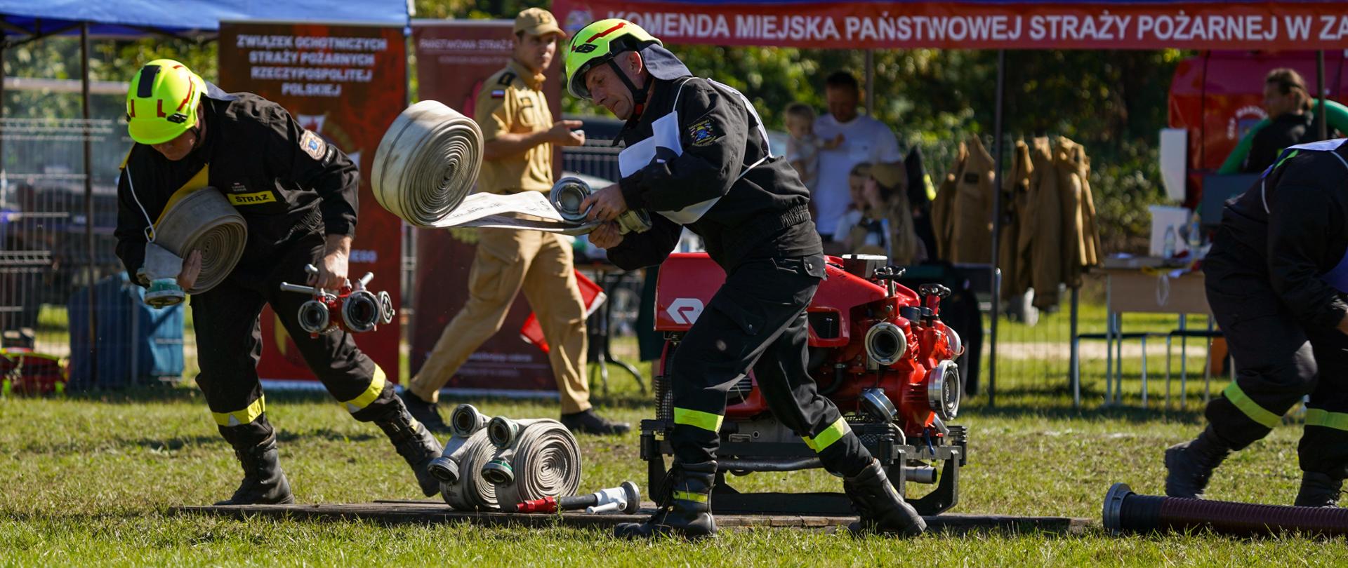 Strażacy wykonują konkurencję - ćwiczenie bojowe. Są w trakcie rozwijania węży. Wszystko dzieje się w biegu. Na zdjęciu widoczna jest ustawiona na podeście pompa, węże pożarnicze. 