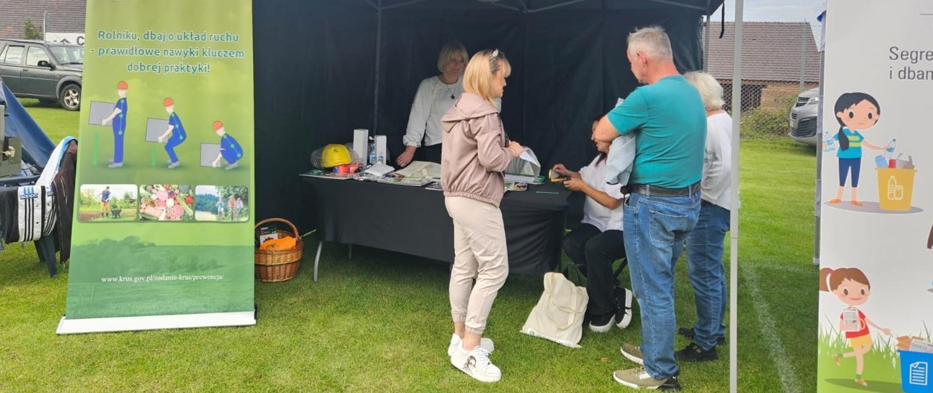 Przy zielonym namiocie stoi grupa osób, a przy stole siedzi na krześle kobieta. Po lewej stronie przed namiotem soi roll-up obrazujący w jaki sposób bezpiecznie podnosić ciężki ładunek z podłogi.