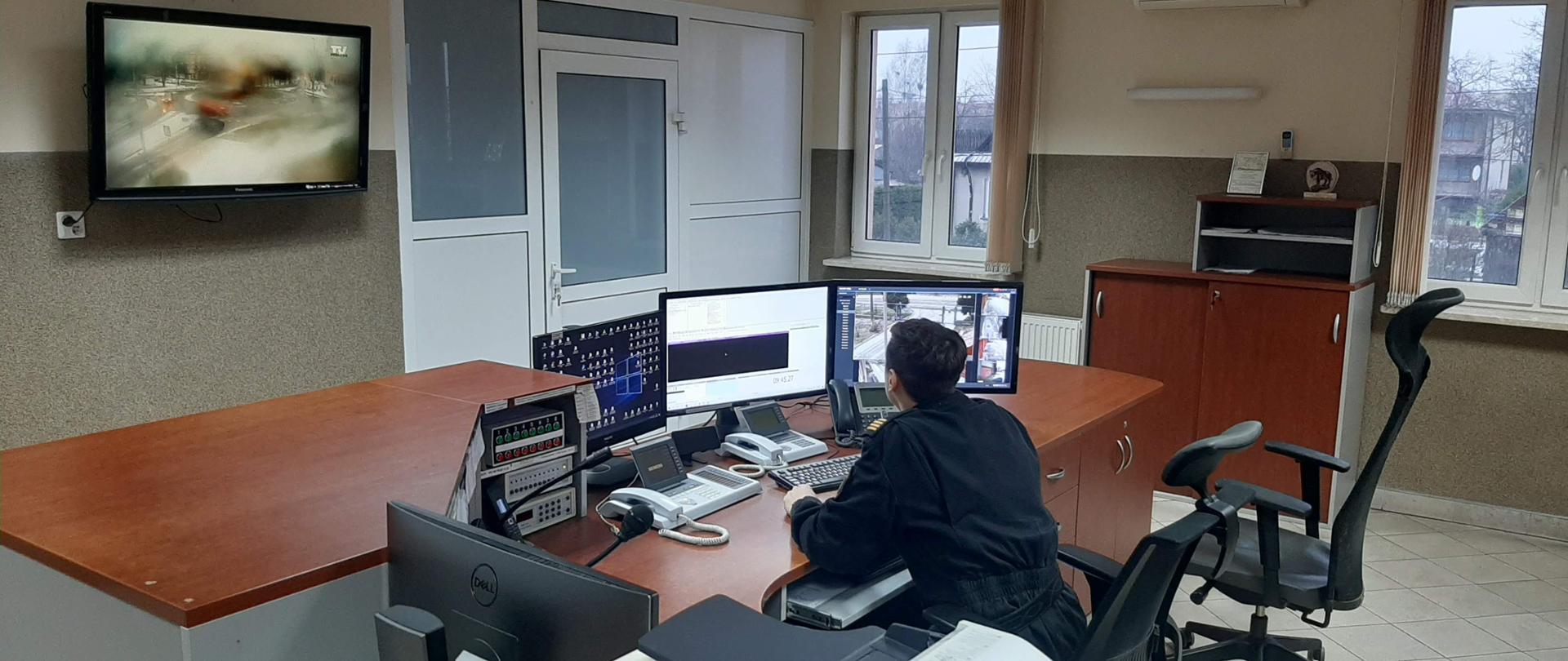 Dyżurny Stanowiska Kierowania Komendanta Powiatowego Państwowej Straży Pożarnej w Hajnówce w czarnym mundurze siedzi przy biurku na którym stoi trzy monitory i trzy telefony.