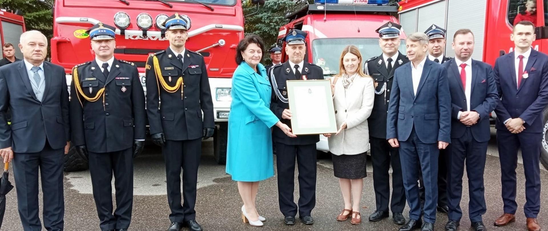 Na zdjęciu widoczni strażacy z OSP i PSP w mundurach galowych koloru granatowego i zaproszeni goście trzymający Decyzję Komendanta Głównego w sprawie włączenia jednostki OSP Rożnowice do KSRG a za nim w tle samochody pożarnicze koloru czerwonego. 