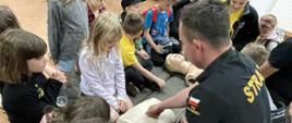 Grupa dzieci oraz strażak prezentujący jak prowadzić resuscytację osoby poszkodowanej na fantomie do ćwiczeń.