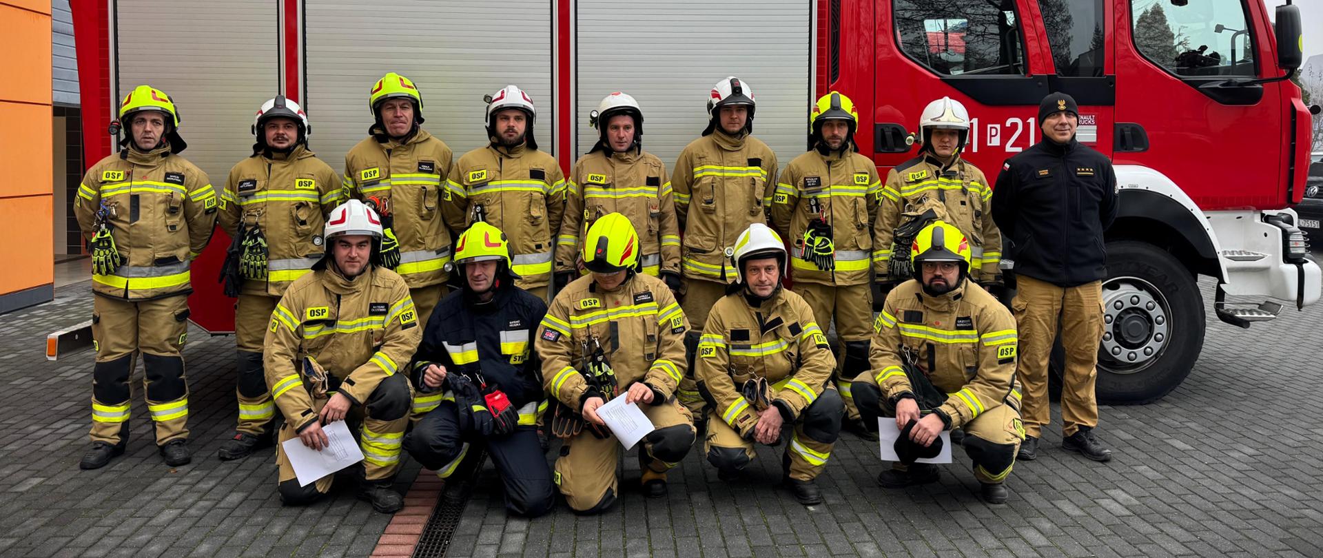 Na zdjęciu znajduje się grupa strażaków w ubraniu specjalnym pozujących do zdjęcia grupowego. Zdjęcie wykonano na utwardzonym parkingu, z widocznymi innymi pojazdami w tle.