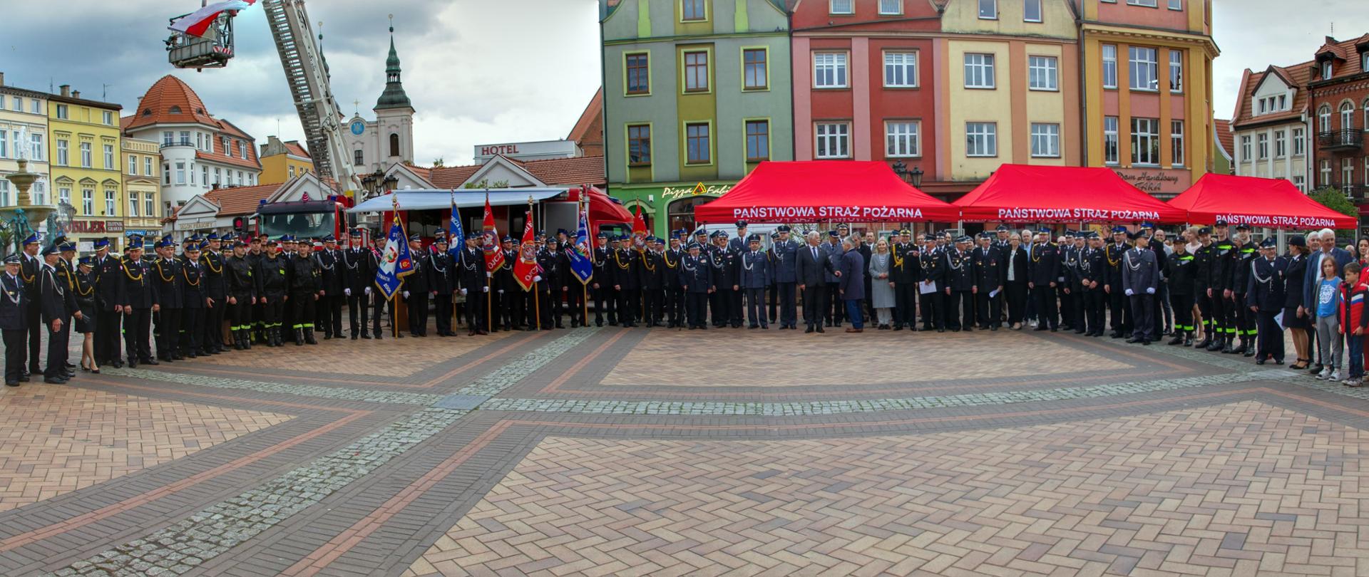 Plac Starego Synku w Chojnicach wyłożony kostką brukową. Zdjęcie grupowe przedawniające uczestników uroczystości, ponad 100 osób, część osób w mundurach. Zdjęcie panoramiczne. 