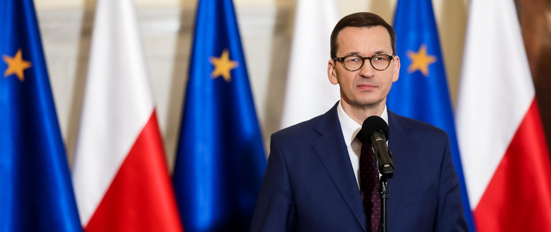 Premier Mateusz Morawiecki stoi przy mikrofonie. W tle flagi Polski i Unii Europejskiej.