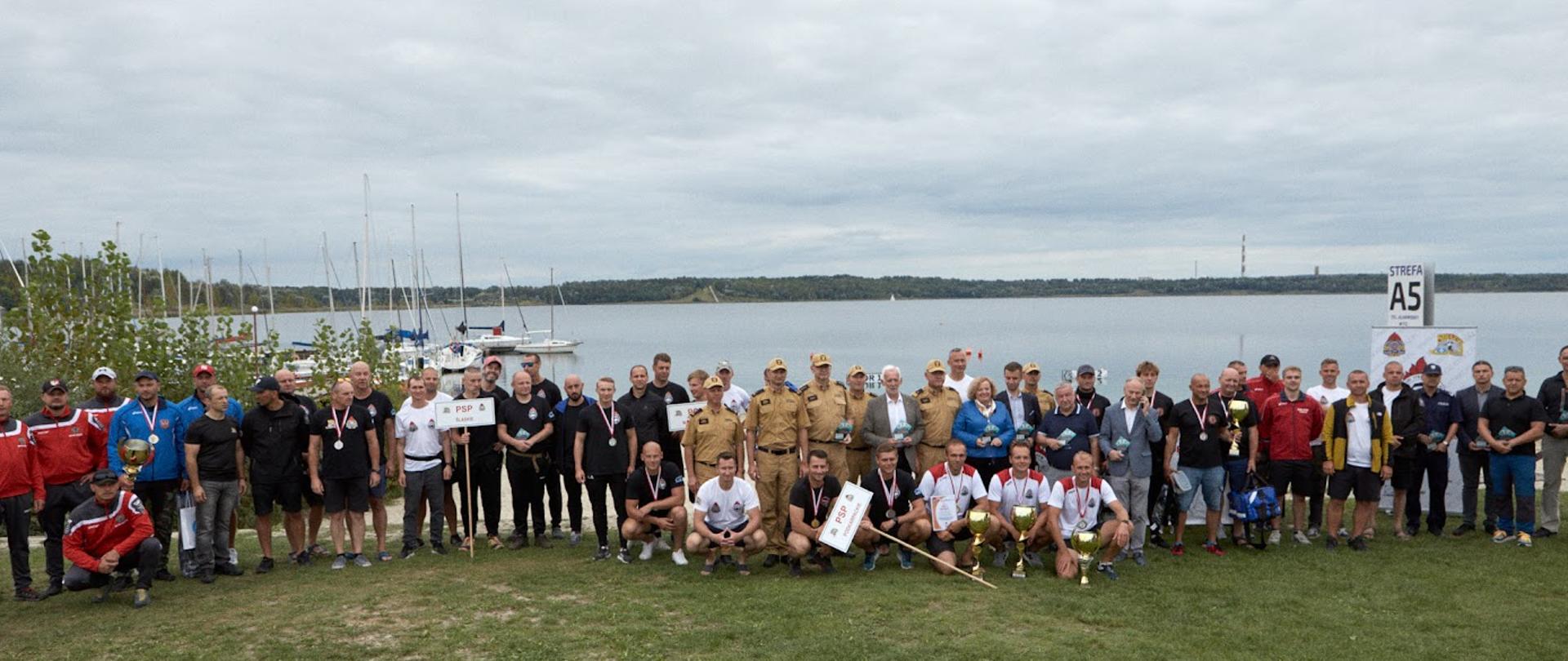 Grupa strażaków i osób cywilnych nad brzegiem jeziora