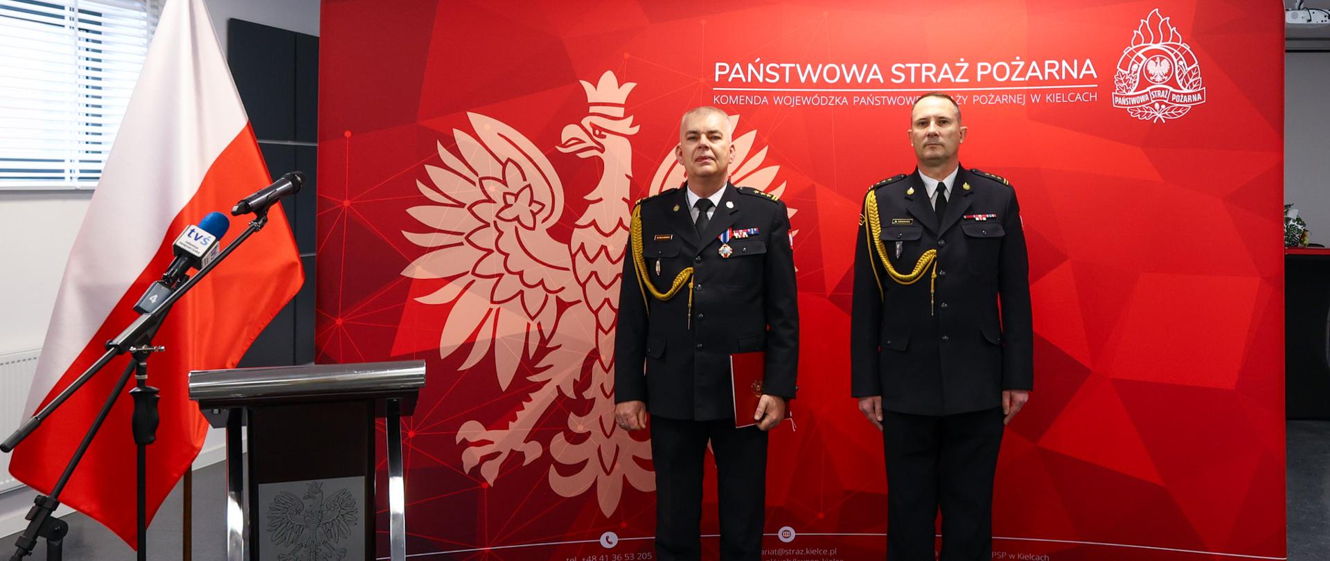 Dwóch oficerów Państwowej Straży Pożarnej w galowych, czarnych mundurach stoi na baczność przed czerwoną ścianką z godłem Polski i napisem „Państwowa Straż Pożarna – Komenda Wojewódzka Państwowej Straży Pożarnej w Kielcach”. Po lewej stronie widoczna jest flaga Polski oraz mównica z mikrofonami. Wnętrze nowoczesnej sali konferencyjnej, jasne oświetlenie, oficjalny charakter uroczystości.