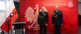 Dwóch oficerów Państwowej Straży Pożarnej w galowych, czarnych mundurach stoi na baczność przed czerwoną ścianką z godłem Polski i napisem „Państwowa Straż Pożarna – Komenda Wojewódzka Państwowej Straży Pożarnej w Kielcach”. Po lewej stronie widoczna jest flaga Polski oraz mównica z mikrofonami. Wnętrze nowoczesnej sali konferencyjnej, jasne oświetlenie, oficjalny charakter uroczystości.