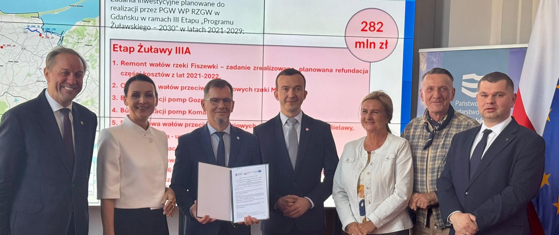 Kilkanaście osób stoi na tle ekranu na którym wyświetlana jest mapka regionu, jedna z tych osób trzyma w rękach podpisaną umowę. Wszyscy są uśmiechnięci i elegancko ubrani. 