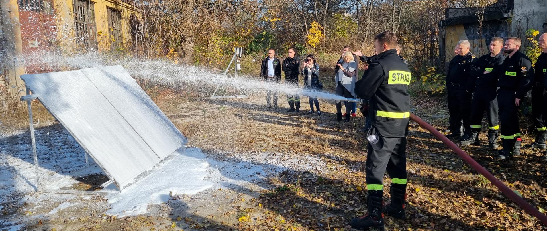 Strażak z wężem pożarniczym podający pianę gaśniczą na panele fotowoltaiczne