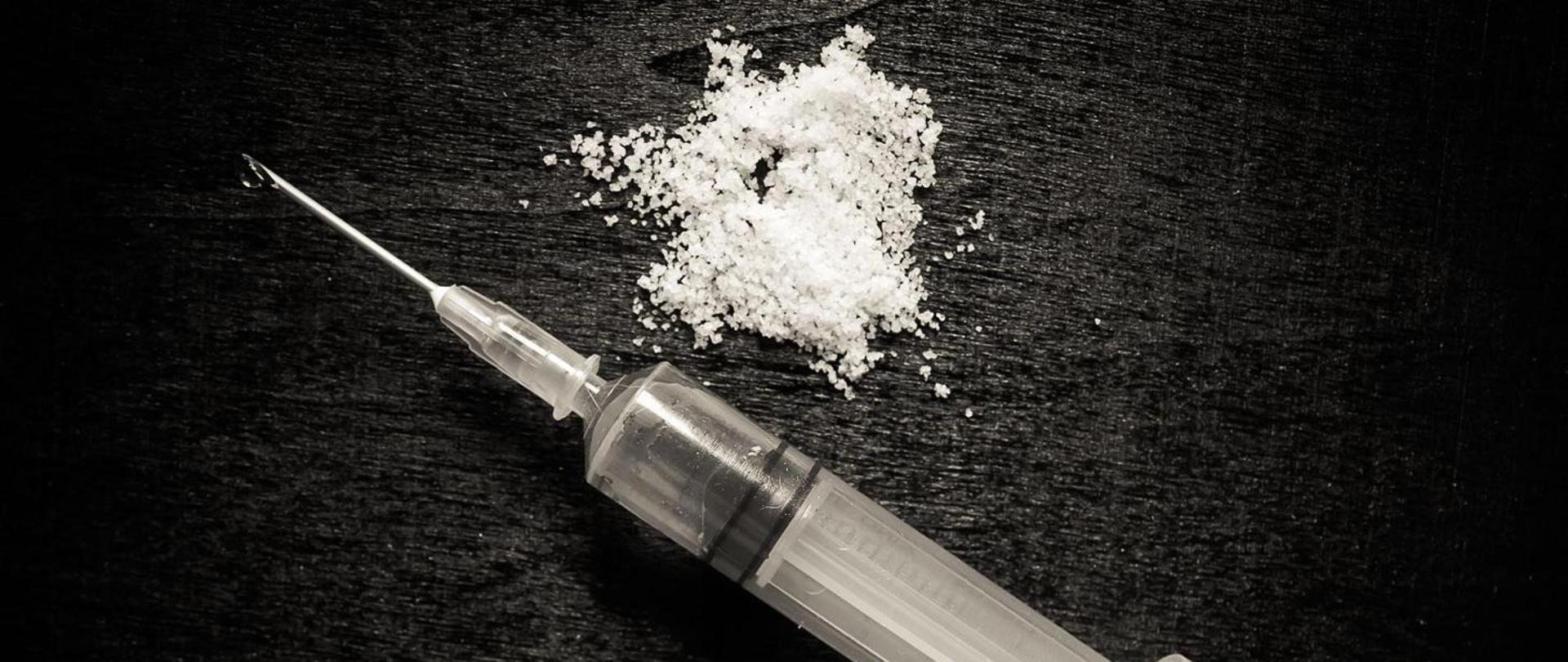 Zdjęcie przedstawia strzykawkę z igłą oraz biały proszek na czarnym tle.