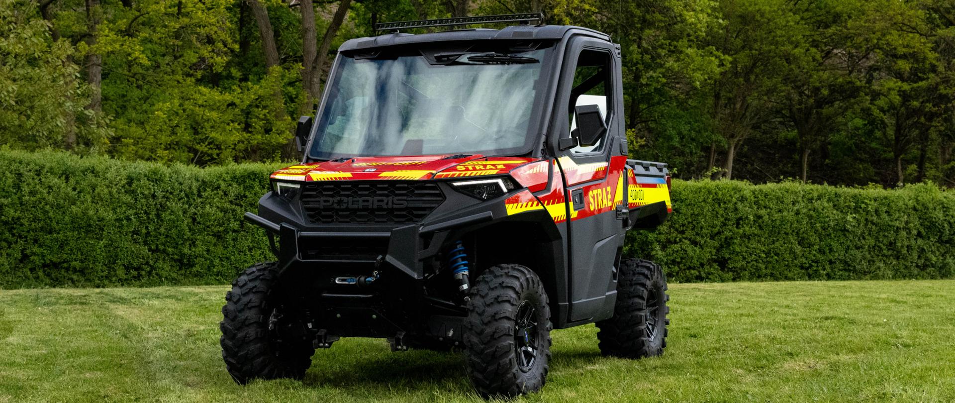 Terenowy pojazd typu UTV (quad z kabiną) marki Polaris Ranger 1000 w jaskrawych barwach strażackich – czerwonej z żółto-neonowymi pasami ostrzegawczymi i napisem „STRAŻ”. Pojazd posiada masywne opony z głębokim bieżnikiem i stoi na zielonej trawie.