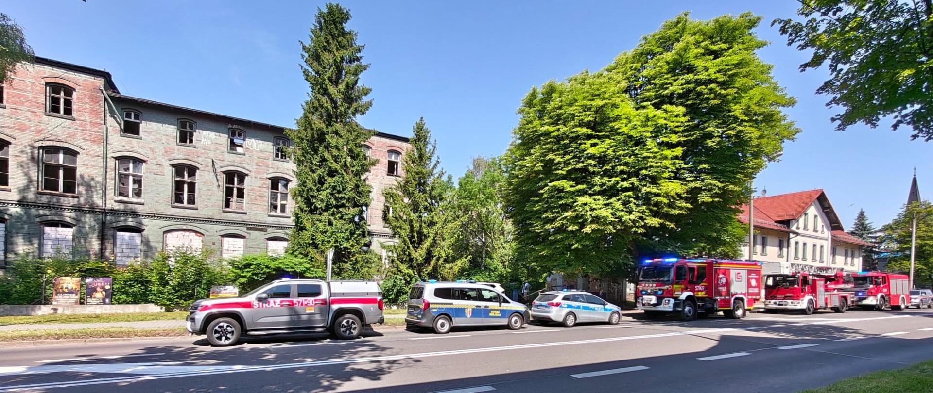 Samochody Państwowej Straży Pożarnej, Policji i Straży Miejskiej stoją na ulicy podczas prowadzonych działań.