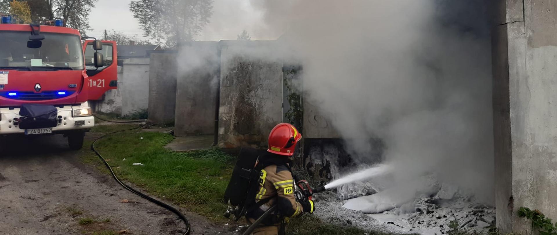 Pomiędzy garażami w zabudowie szeregowej pali się jeden z nich. Na zdjęciu widać kucniętego strażaka który gasi wnętrze palącego się garażu. W oddali na drodze stoi strażacki samochód.