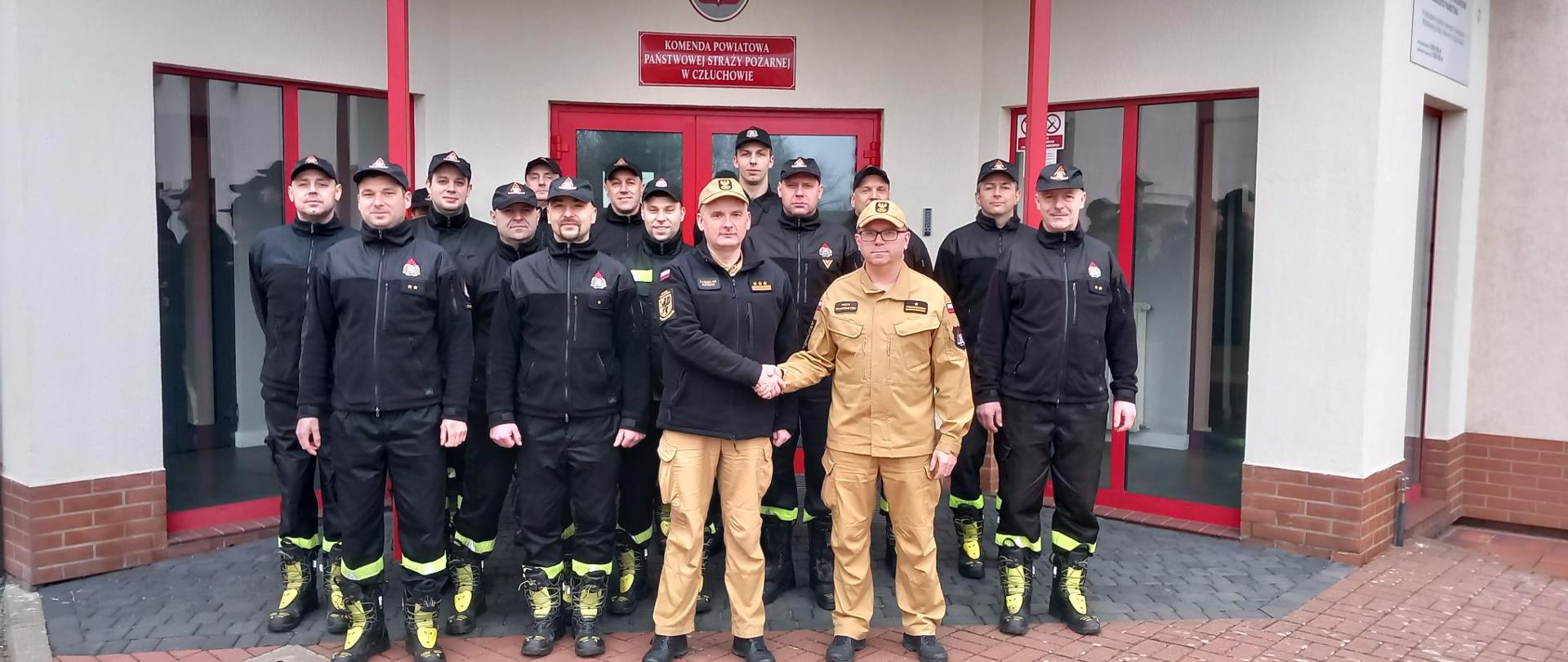 Zdjęcie grupowe po uroczystej zmianie służby. Komendant Powiatowy Państwowej Straży Pożarnej w Człuchowie st. bryg. Radosław Batruch gratuluje mł.bryg. Piotrowi Frankenstein awansu na stanowisko Dowódcy Jednostki Ratowniczo – Gaśniczej w Komendzie Powiatowej Państwowej Straży Pożarnej w Człuchowie. Obok na tle budynku komendy stoją funkcjonariusze ze zmian służbowych.