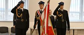 Poczet sztandarowy stoi oddając honory za nimi stoi strażak kobieta w mundurze wyjściowym ze sznurem oraz ustawione są krzesła oraz biurka.