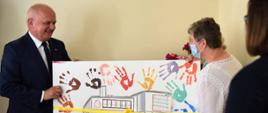 Wojewoda wraz z jedną z seniorek trzyma duży obraz pokazujący szkic budynku domu seniora, podpisem Dzienny Dom Senior + w Nowej Soli oraz kolorowymi, odbitymi dłońmi seniorów uczęszczających na zajęcia. Obraz jest przewiązany żółtą wstążką 