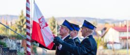 Zdjęcie przedstawia poczet flagowy (3 strażaków) podczas podnoszenia flagi na maszt. Zdjęcie wykonane na zewnątrz.