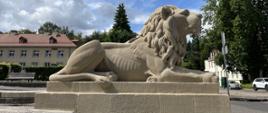 Na zdjęciu widoczna jest kamienna rzeźba lwa leżącego na prostokątnym cokole. Lew spogląda w prawą stronę, jego grzywa jest bujna i szczegółowo wyrzeźbiona, a na ciele widać zarysowane żebra. Pomnik znajduje się na otwartym placu, w tle rosną wysokie drzewa oraz stoją jasne budynki z czerwonymi dachami i białymi oknami. Niebo jest częściowo zachmurzone, między chmurami przebija błękit. Po lewej stronie widoczne są ławki i niska zieleń, po prawej – zaparkowane auta i znak drogowy.