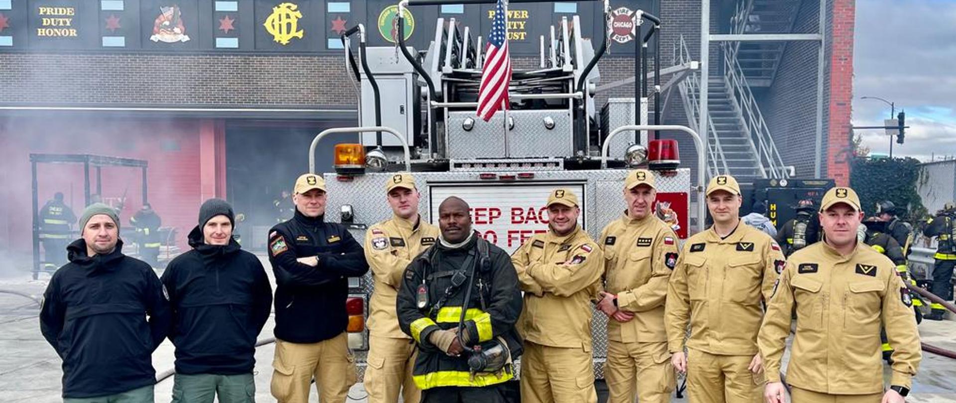 Polska delegacja pozuje do zdjęcia z amerykańskim czarnoskórym strażakiem na tle samochodu strażackiego z flagą USA przed budynkiem Akademii Pożarniczej