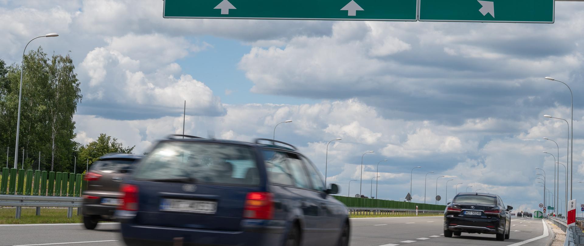 Zdjęcie przedstawia samochody jadące dwupasmową drogą ekspresową. Dwa skręcają w prawo do zjazdu z drogi.