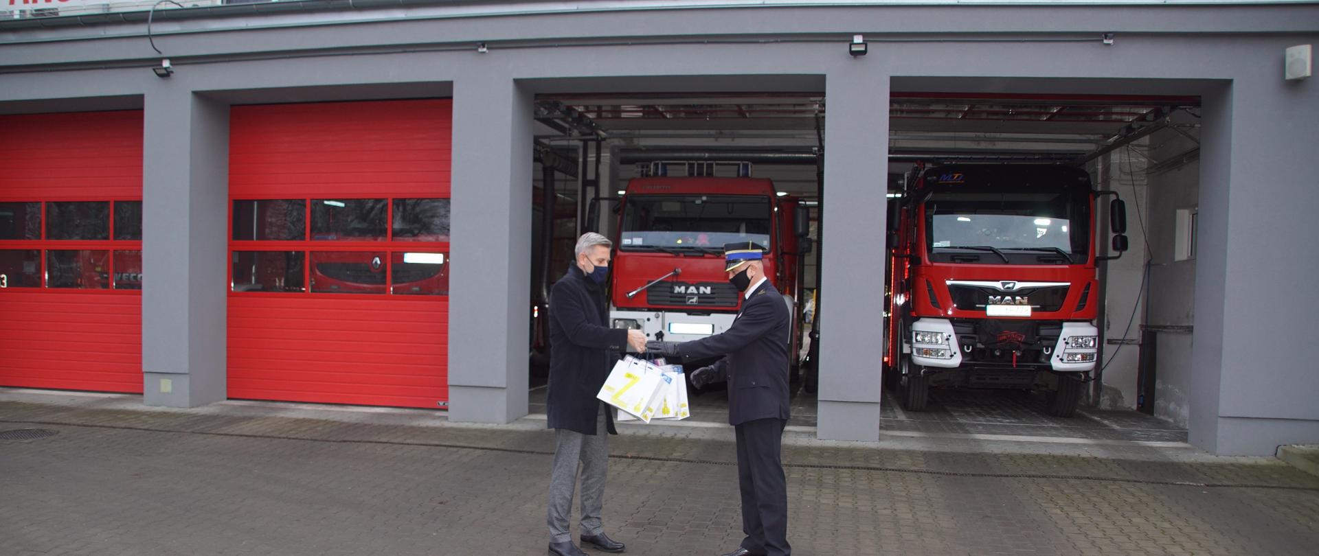 Prezydent Miasta Zgierza Przemysław Staniszewski przekazuje czujki Komendantowi KP PSP w Zgierzu, na tle czerwonych bram wyjazdowych oraz samochodów ratowniczo gaśniczych marki MAN z Jednostki Ratowniczo Gaśniczej w Zgierzu.