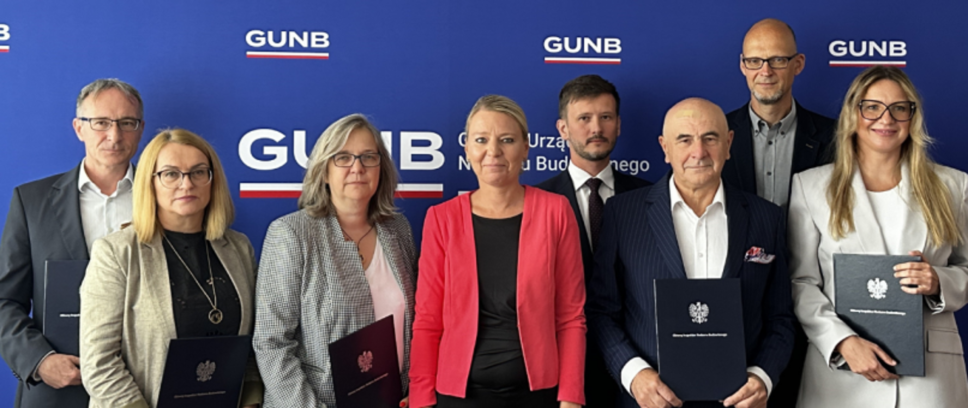 Grupa osób stojąca przed ścianką GUNB z teczkami w dłoniach, z widocznym logo Głównego Urzędu Nadzoru Budowlanego.