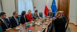 W sali przy podłużnym stole siedzą ludzie, u szczytu stołu minister Wieczorek mówi, za nim pod ścianą flagi Polski i UE.