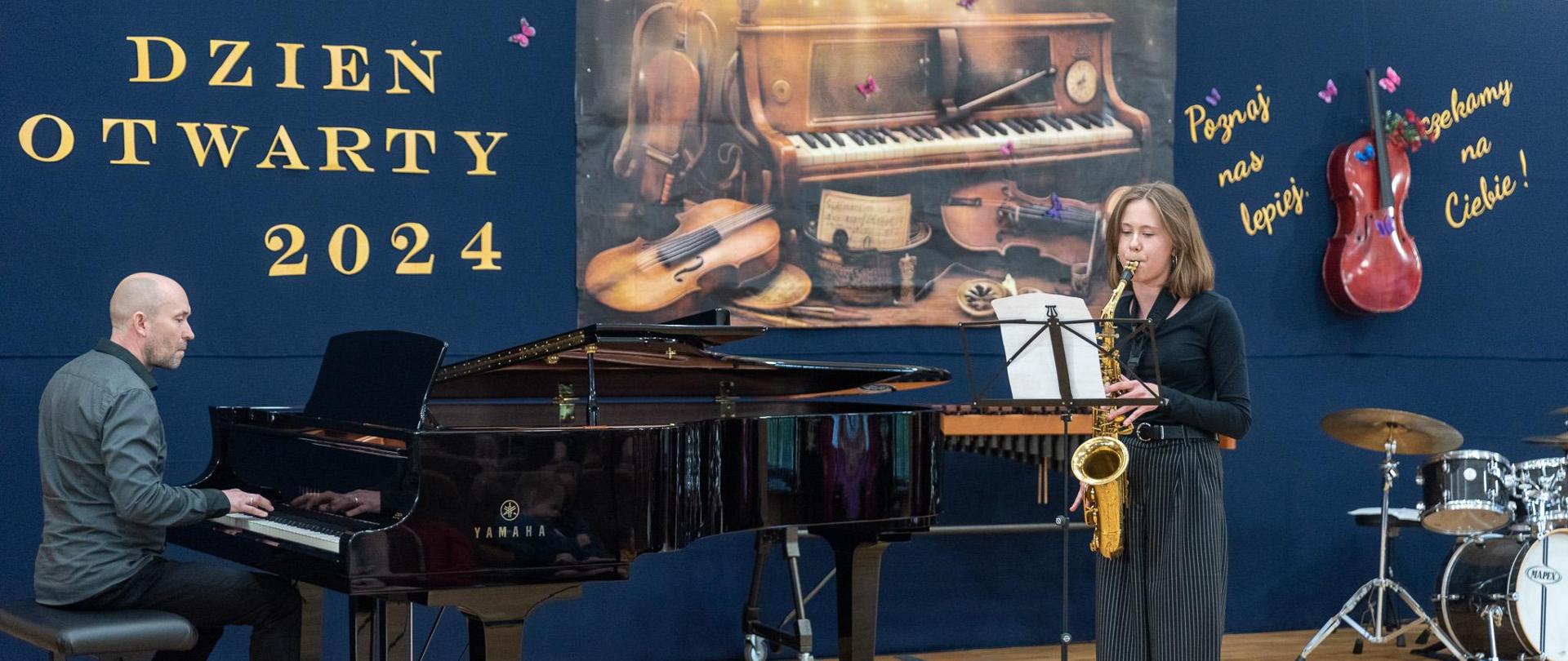 Na scenie saksofonistka gra i pianista jej akompaniuje. W tle napis "Dzień Otwarty 2024" i wisząca wiolonczela na ścianie.