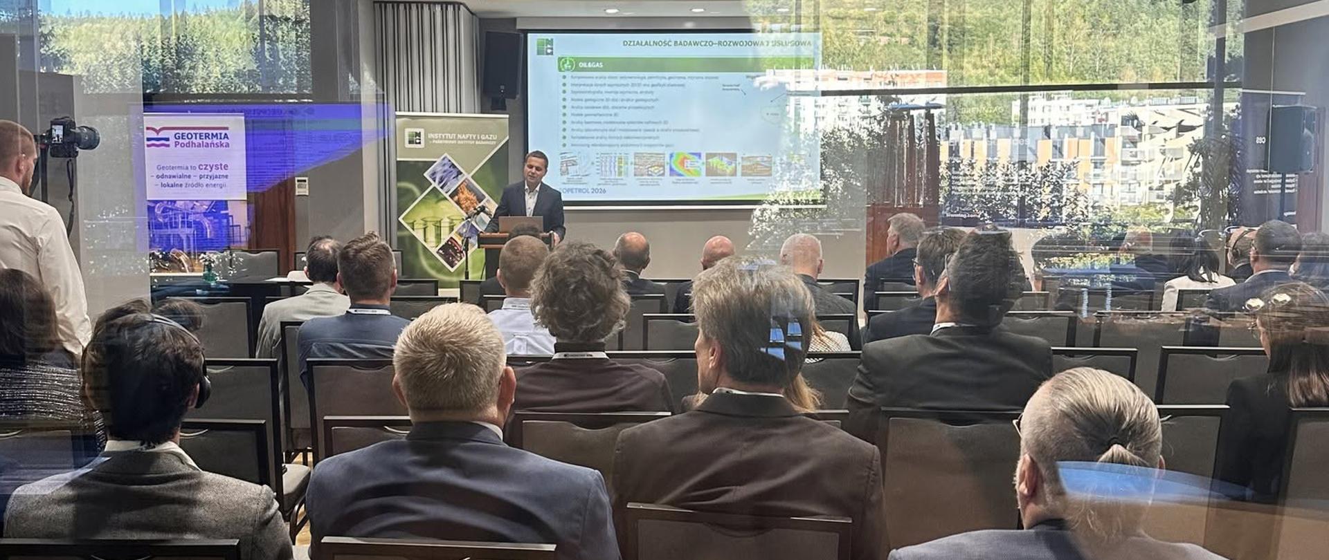 Konferencja geologiczna z udziałem uczestników. Na pierwszym planie widoczna jest publiczność, a w tle prelegent prowadzi prezentację dotyczącą działalności badawczo-rozwojowej i przemysłowej.