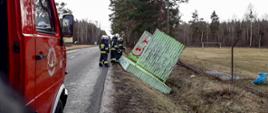 W rowie przy drodze asfaltowej leży przewrócona wiata. W tle widać drzewa oraz kabinę samochodu pożarniczego.