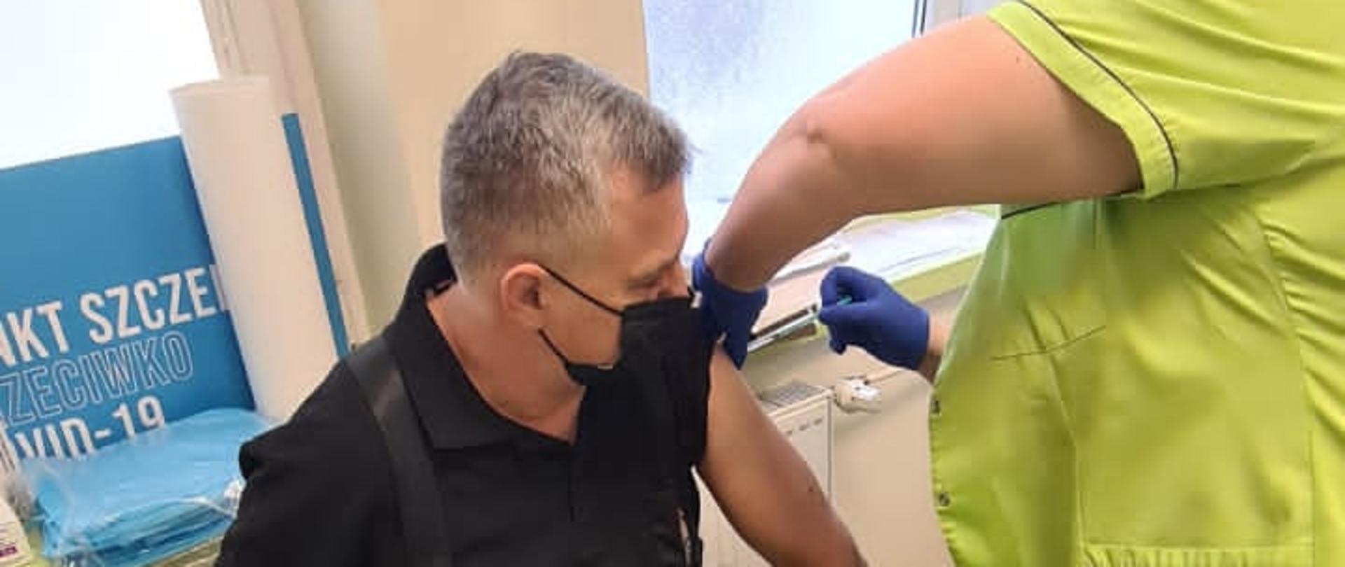 Na zdjęciu widać strażaka siedzącego na kozetce. Jest to kujawsko-pomorski komendant wojewódzki PSP. Strażak ma podwinięty rękaw, a obok niego stoi kobieta wbijająca w jego ramie igłę ze strzykawką. Zdjęcie wykonano w punkcie szczepień.