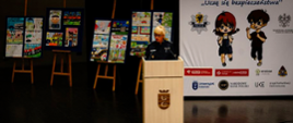 Policjantka przemawia przy mównicy za nimi na sztalugach ułożone są prace plastyczne oraz baner akcji Uczę się bezpieczeństwa" z logo Policji oraz podmiotów partnerów wspierających akcję. 