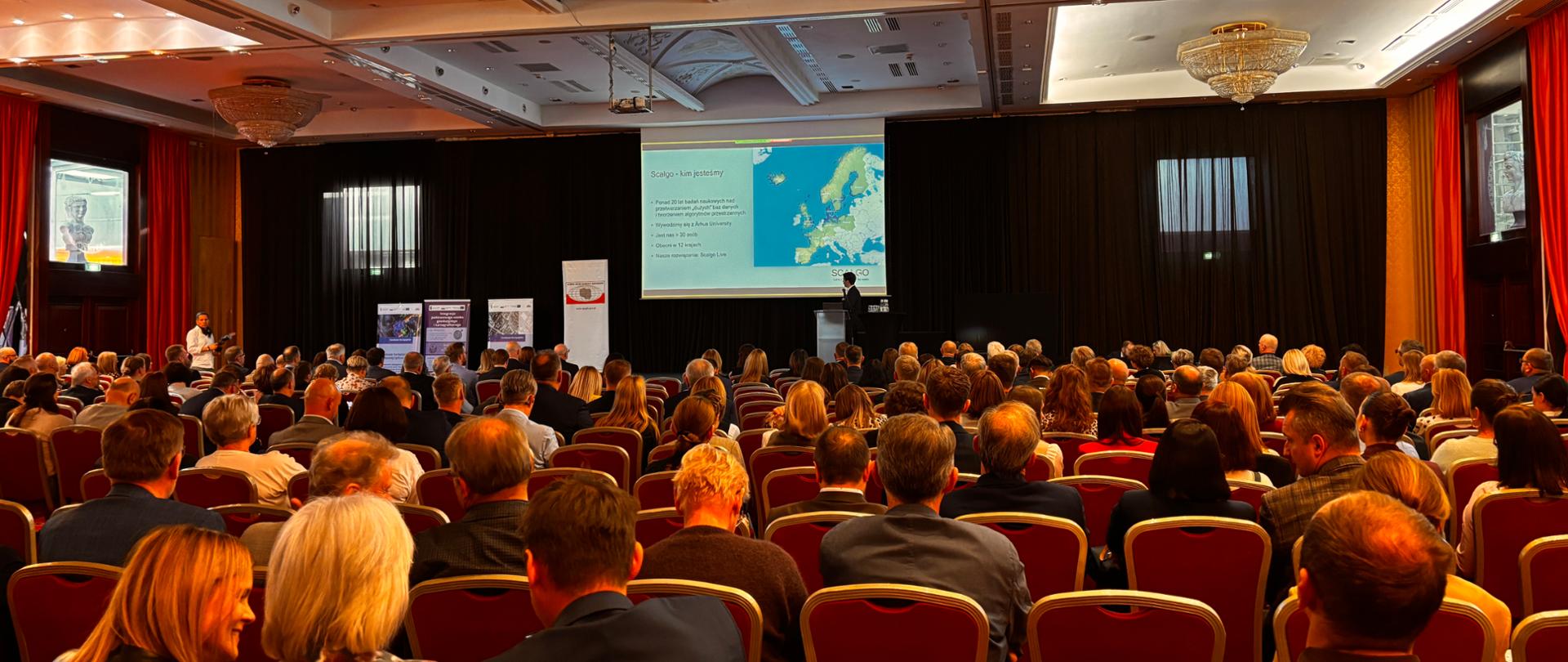 Konferencja Służby Geodezyjnej i Kartograficznej. Na dużej sali konferencyjnej uczestnicy. W tle ekran i prelegent prowadzący panel merytoryczny