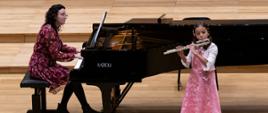 Dziewczynka stojąc na środku estrady sali koncertowej PSM gra na flecie, za nią na fortepianie gra kobieta.