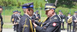 Na zdjęciu widoczni Małopolski Komendant Wojewódzki PSP wręczający oficerowi dowódcy uroczystości odznaczenie. Komendant w galowym granatowym mundurze i w czapce rogatywce. Strażak odbierający odznaczenie w galowym granatowym mundurze i w czapce rogatywce.