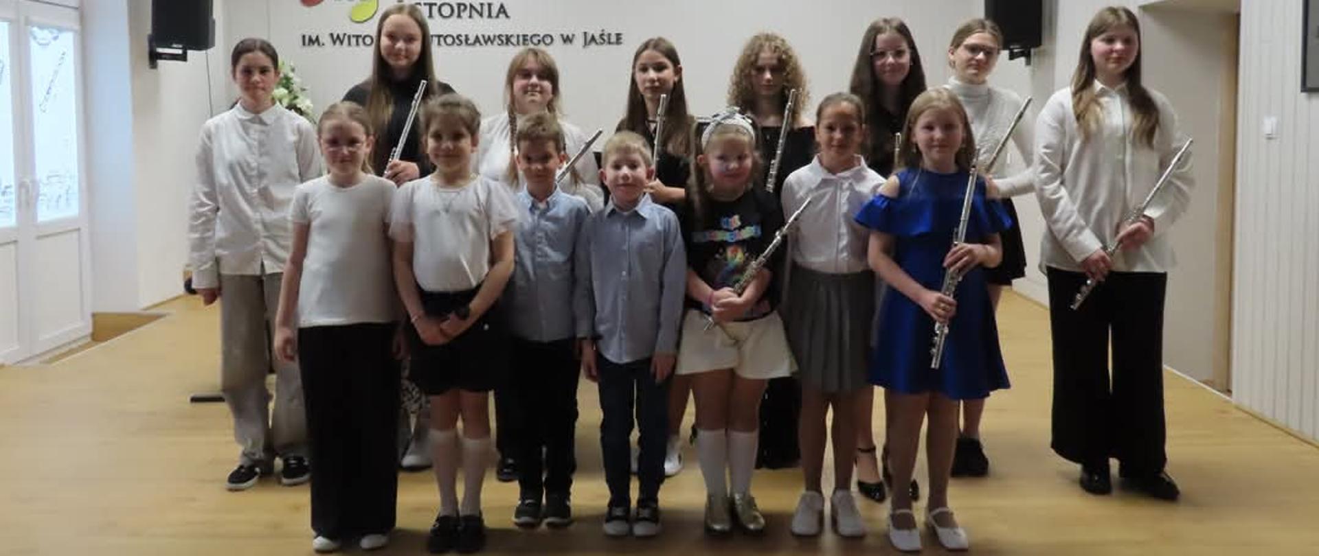 Grupowe zdjęcie uczniów z Państwowej Szkoły Muzycznej I stopnia im. Witolda Lutosławskiego w Jaśle, wykonane na scenie szkolnej sali koncertowej. Uczniowie trzymają flety, uśmiechają się do obiektywu i są ubrani odświętnie. W tle widoczna jest nazwa szkoły na ścianie. Kolorystyka zdjęcia jest stonowana i naturalna, z przewagą jasnych, neutralnych barw – dominują biel, beże i pastele, co tworzy spokojną, elegancką atmosferę i podkreśla formalny charakter wydarzenia.