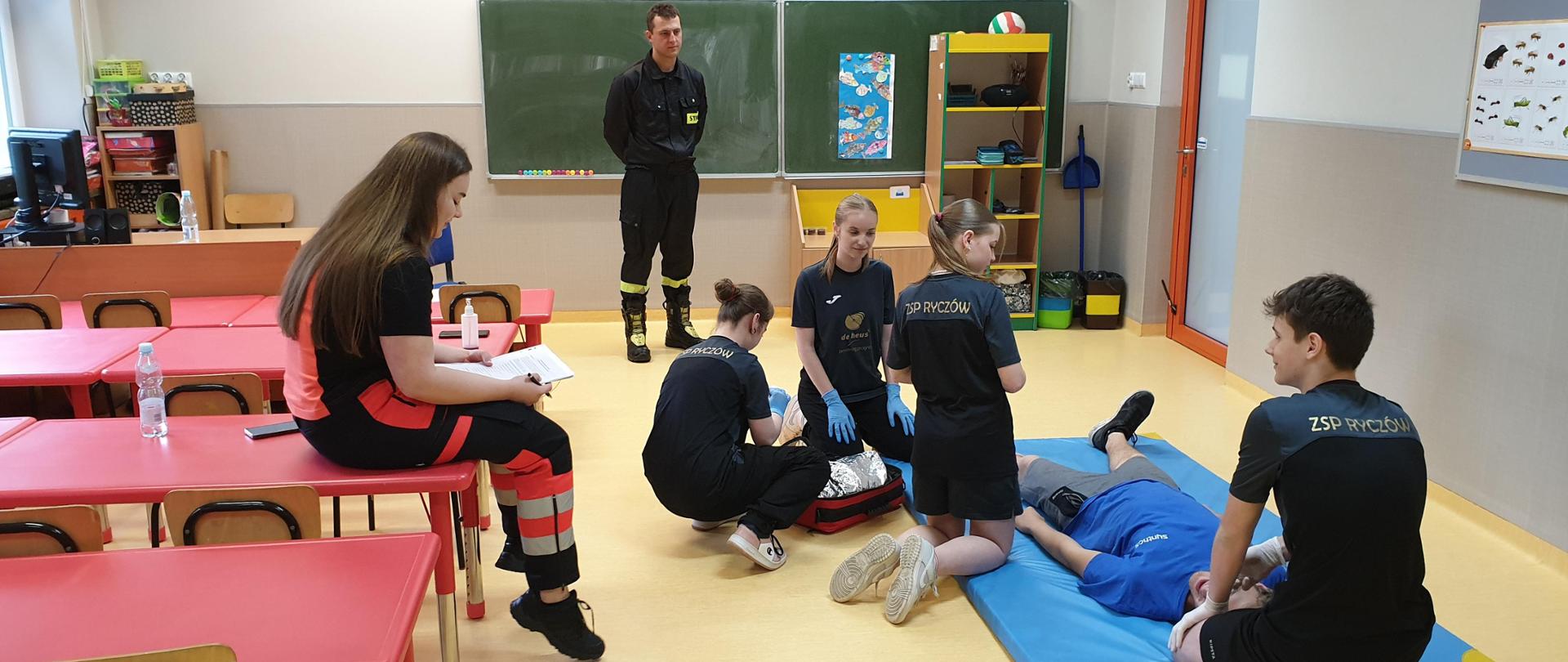 Widok z tyłu. Scenka-zadanie podczas turnieju. Młodzież udziela pierwszej pomocy osobie poszkodowanej w sali lekcyjnej. Zadanie ocenia strażak i ratowniczka medyczna.