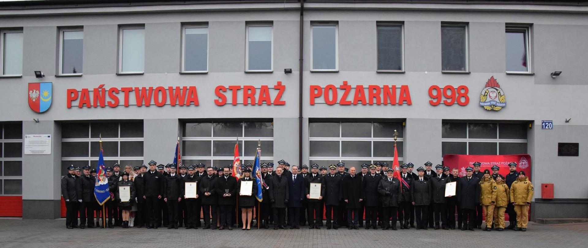 Duża grupa strażaków w mundurach galowych stoi w szeregu przed budynkiem Komendy Powiatowej PSP. Przed nimi widoczne są sztandary oraz osoby trzymające dyplomy. Nad bramami garażowymi znajduje się napis „Państwowa Straż Pożarna 998”.