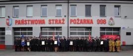 Duża grupa strażaków w mundurach galowych stoi w szeregu przed budynkiem Komendy Powiatowej PSP. Przed nimi widoczne są sztandary oraz osoby trzymające dyplomy. Nad bramami garażowymi znajduje się napis „Państwowa Straż Pożarna 998”.