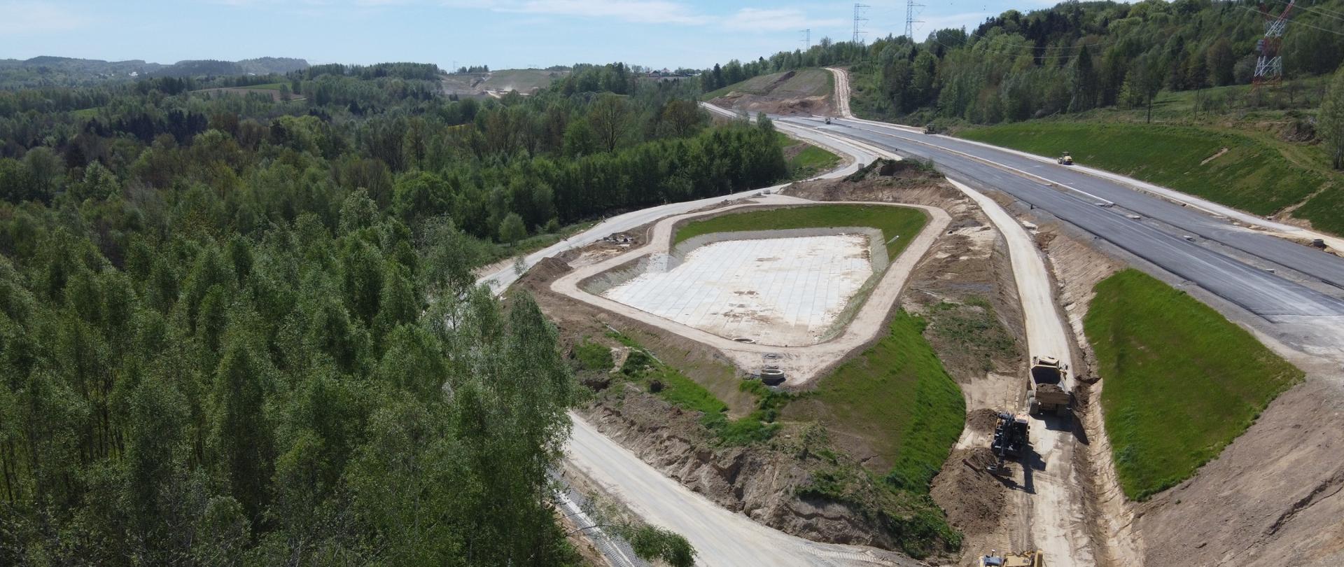 Zdjęcie z drona pokazuje teren budowy drogi wśród zielonych wzgórz i lasów. W centrum znajduje się pusty, betonowy zbiornik o prostokątnym kształcie, otoczony drogą techniczną. Po prawej biegnie nowa jezdnia asfaltowa, a na dole pracują maszyny budowlane. W tle widać lasy, pola i słupy energetyczne pod jasnym niebem.