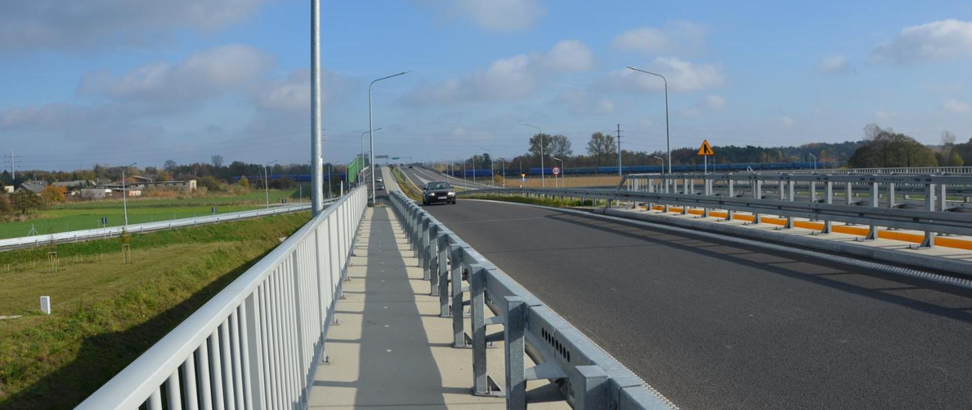 Słoneczny dzień, na niebie lekkie chmury. Widok na jezdnię, w tle widoczny zbliżający się samochód.