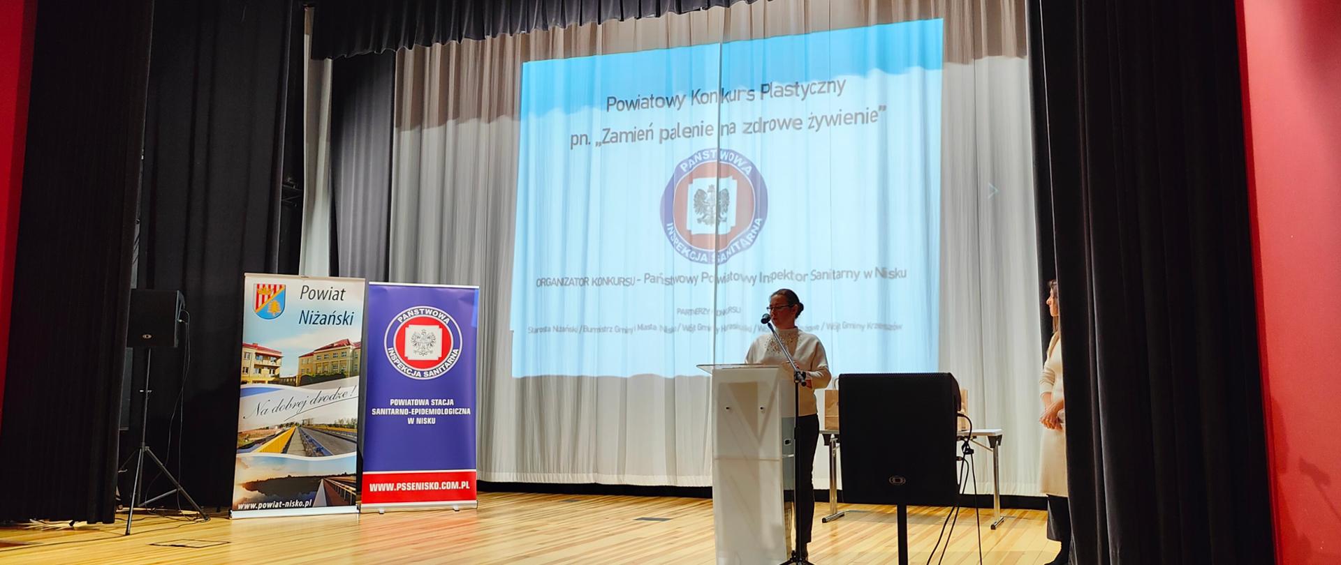 PPIS w Nisku - Pani Magdalena Rachwał wygłaszająca przemówienie, w tle wyświetlony slajd z napisem "Powiatowy Konkurs Plastyczny pn. "Zmień Palenie na Zdrowie Żywienie" i logo Inspekcji Sanitarnej