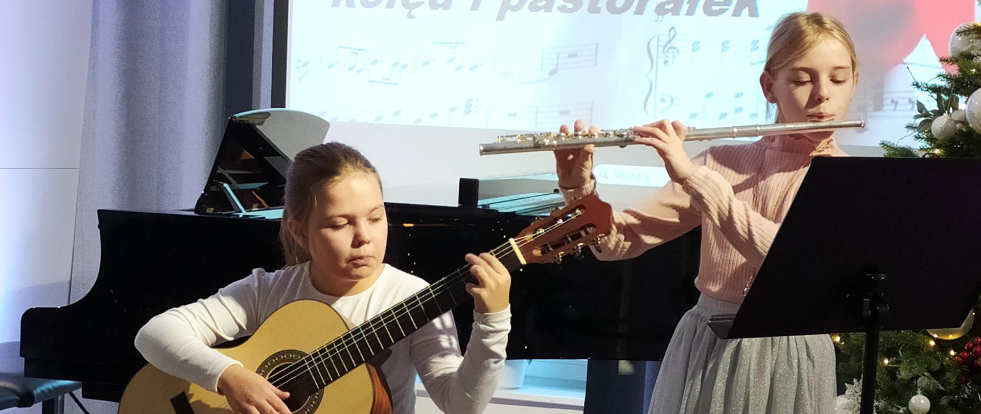 Uczennice grające w duecie, jedna na gitarze, a druga na flecie. Z prawej strony stoi ubrana choinka, a z lewej z tyłu fortepian. W tle wyświetlony jest slajd z nutkami i nazwą koncertu. 