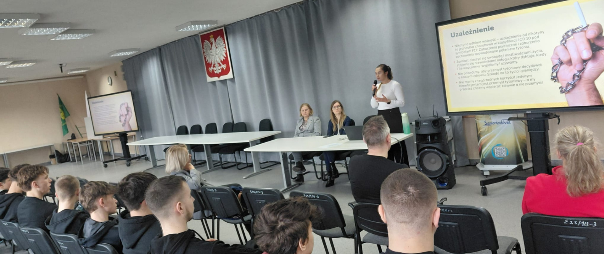 Widok z sali konferencyjnej, osoby prowadzące wystąpienie oraz uczniowie siedzący na krzesłach.