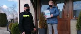 Zdjęcie przedstawia strażaka OSP oraz kobietę, która znajduje się na schodach przed wejściem do budynku. Kobieta trzyma w rękach ulotkę dotyczącą szczepień przeciwko COVID-19. Każda z osób ma na twarzy maseczkę chirurgiczną. Wokół tych osób znajdują się zielone tuje. 